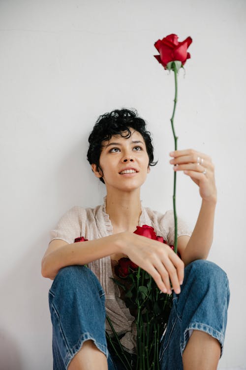 Glad female with short hair looking at red flower in hand while sitting near white background with bouquet of roses