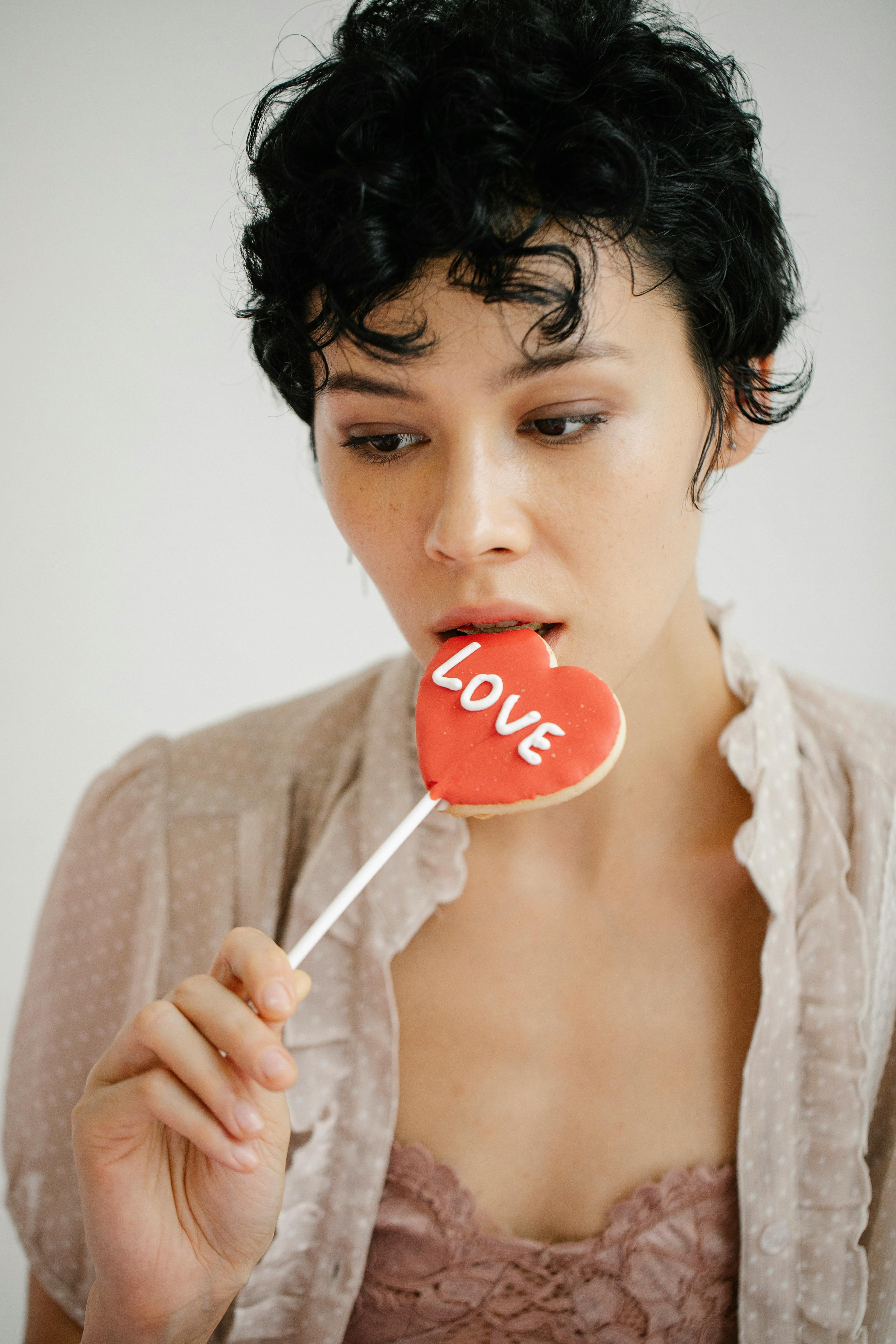 Thoughtful female eating sweet red cookie with Love inscription during Saint Valentines Day celebration on white background in light studio studio