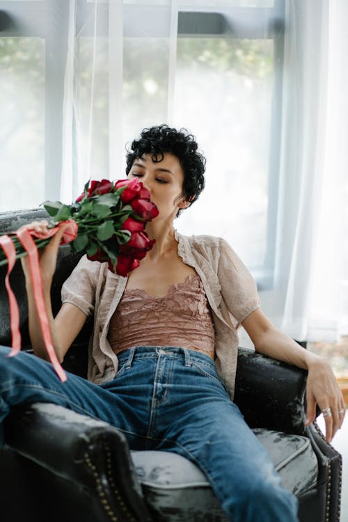 Relaxed female in casual clothes with bouquet of red flowers in hand sitting in comfortable armchair near window in room