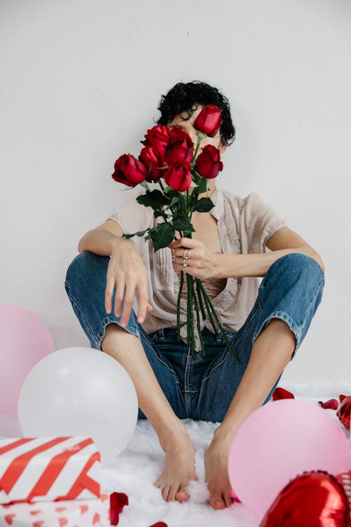 Full body of unrecognizable barefoot female with bouquet roses in hand sitting near white wall amidst colorful balloons and gift boxes during Saint Valentines day