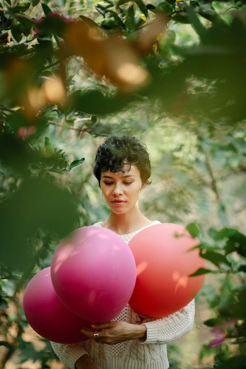 Pensive female looking at colorful balloons in hands while standing among green trees during festive occasion on blurred background