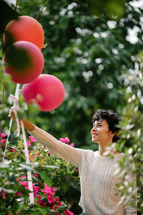 Side view of happy female with colorful balloons in outstretched arm standing standing near blooming bushes on blurred background during festive occasion