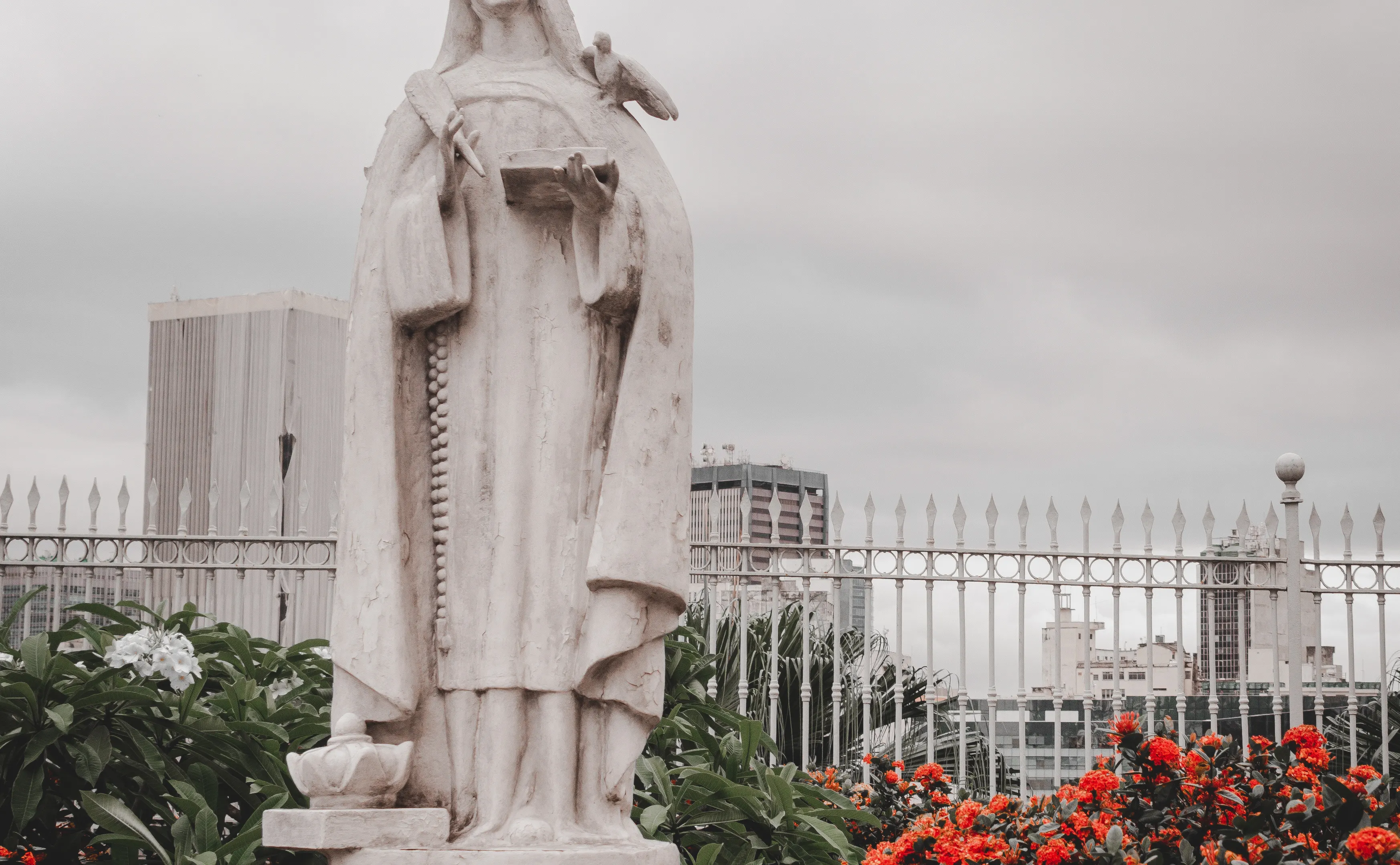 Marble statue of Santa Teresa de Jesus amidst city buildings and vibrant flowers.