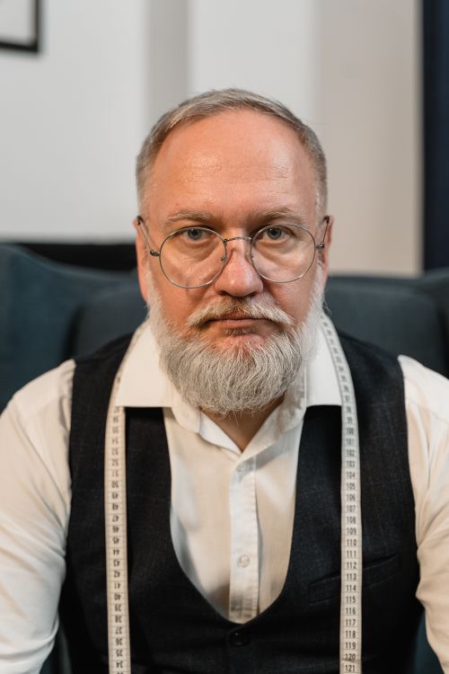 Free Senior male tailor with glasses and white beard wearing a dress shirt and vest, sitting indoors. Stock Photo