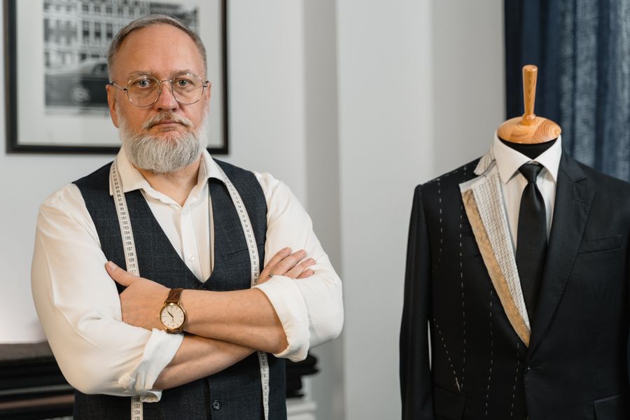 Senior tailor standing with arms crossed beside a bespoke suit on a mannequin in a stylish atelier.