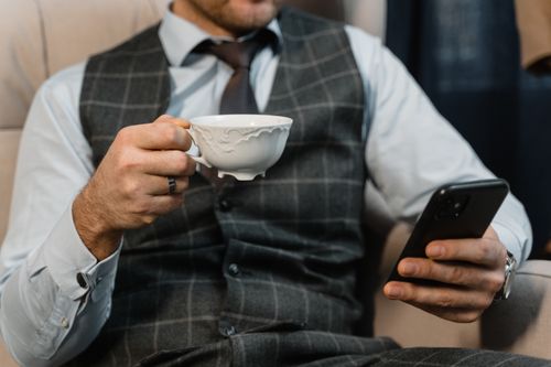 Free A well-dressed businessman in a checkered suit holds a smartphone and teacup, showcasing modern elegance. Stock Photo