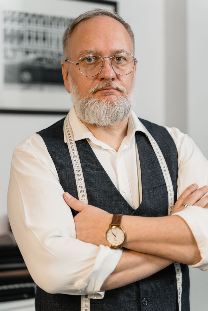 A confident tailor with eyeglasses and a white beard poses with folded arms in a workshop.
