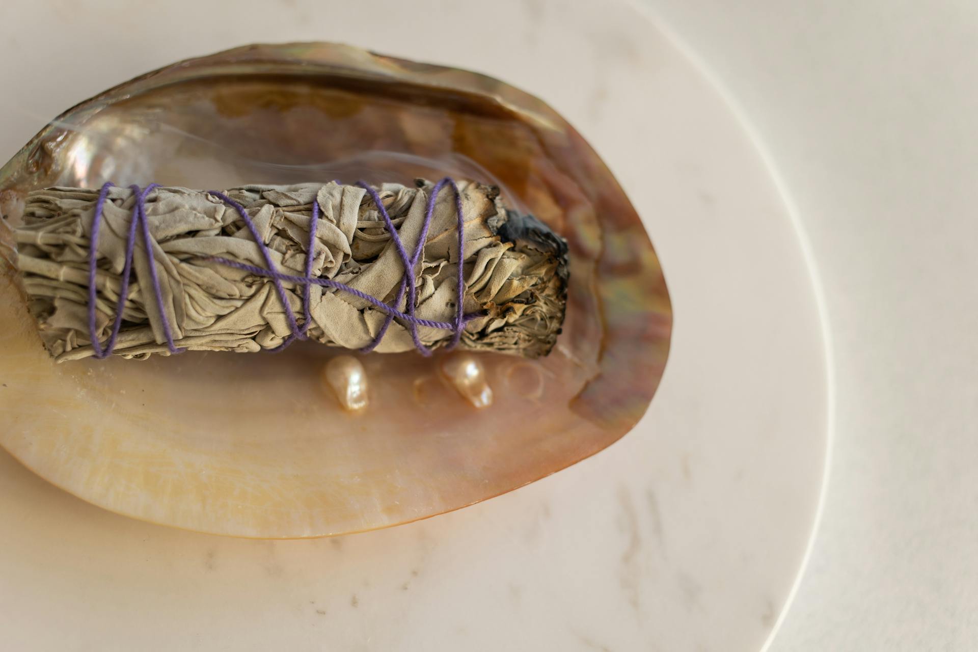 A sage smudge stick wrapped in violet thread rests in a shell with pearls, creating a calming still life setting.