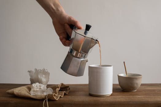 A hand pouring coffee from a moka pot into a ceramic cup beside crystals on a table.