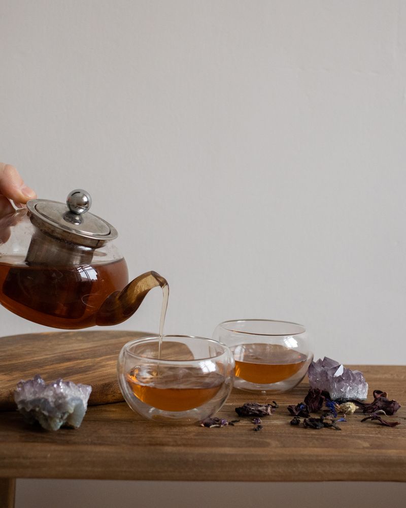 手持玻璃茶壺將熱茶倒入茶杯,木托盤上擺放紫水晶原礦