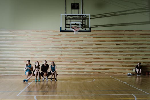 Young female basketball players sitting on a gym bench under a basketball hoop, resting post-practice.