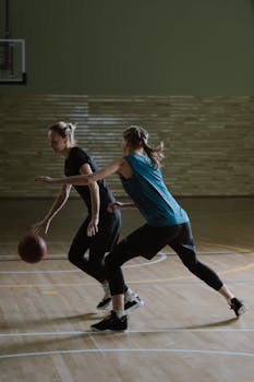 Two women play a competitive basketball game on an indoor court, showcasing teamwork.