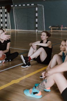 A group of young women athletes sitting on a basketball court discussing strategy indoors.