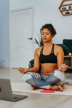 A woman practicing yoga and meditation at home using an online class on her laptop.
