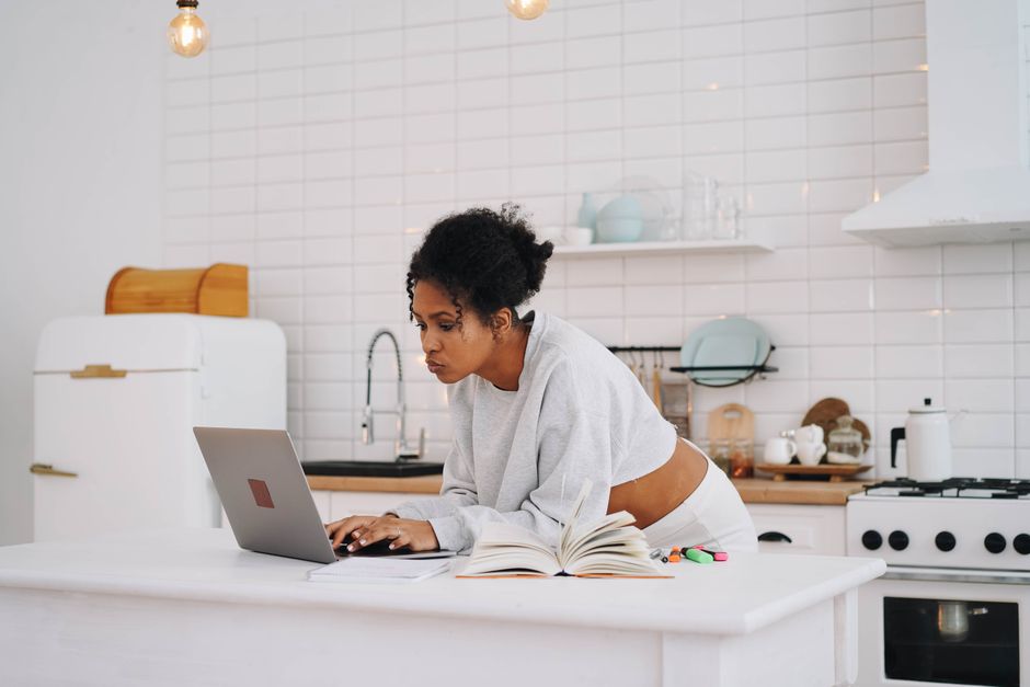 A woman with afro hair works on her laptop in a stylish kitchen setting.