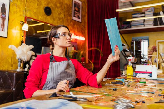 A woman artist in a vibrant studio holding a painted board, surrounded by tools and art supplies.