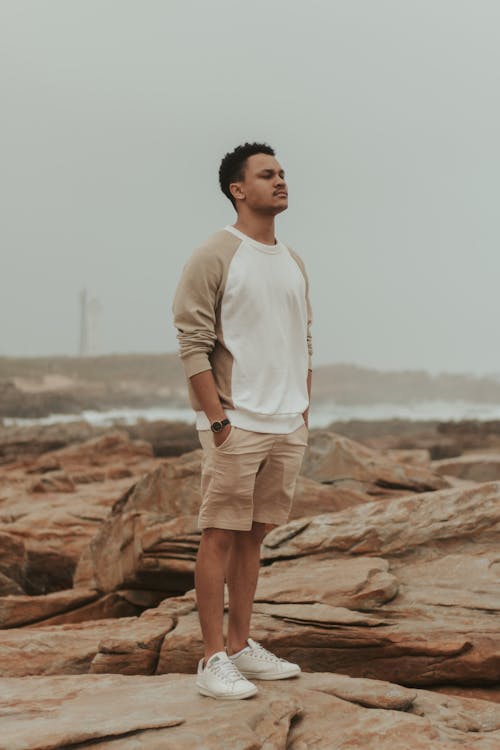 Full body of African American male in casual clothes standing on stony coast near waving river in cloudy day