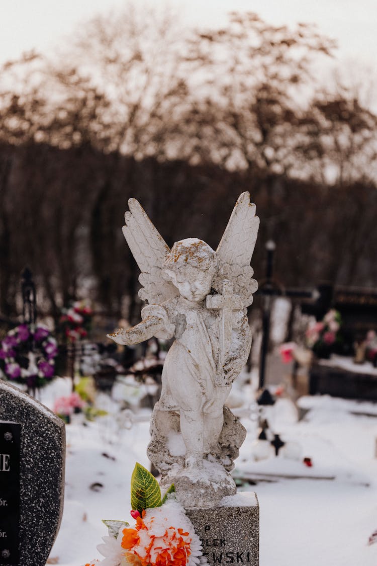 A Snow Covered Angel Statue Beside Snow Covered Flower On A Grave