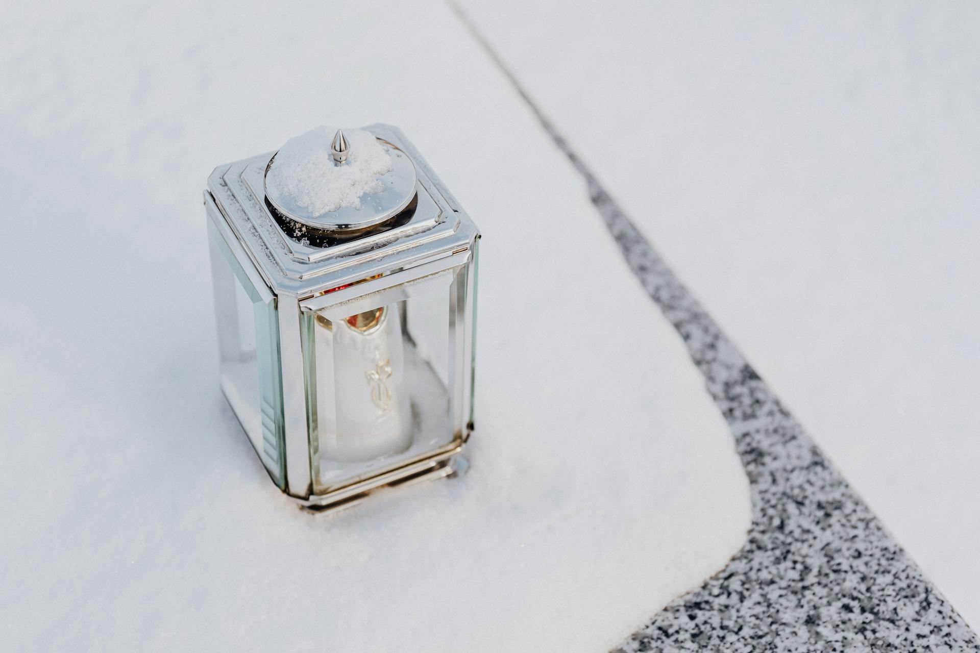 A glass lantern with a candle inside, resting on a snowy stone surface.