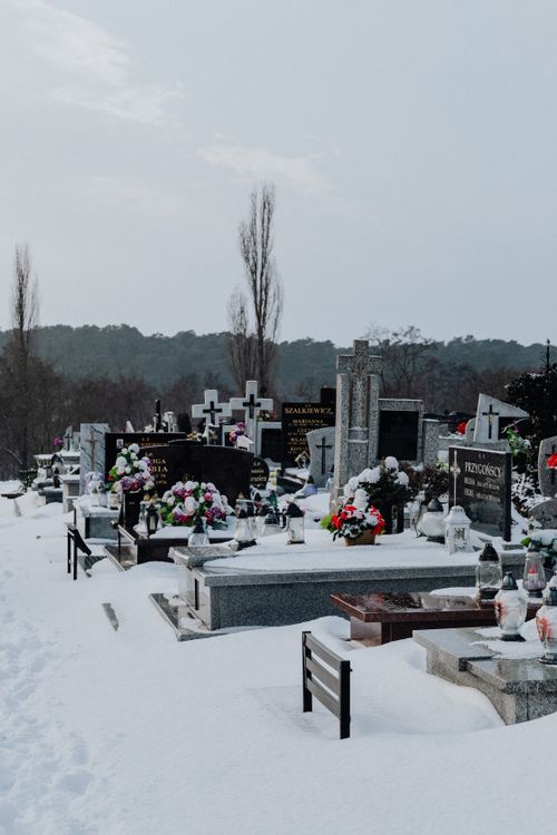 Free A serene winter scene of a snow-covered cemetery with tombstones and floral tributes under a soft, overcast sky. Stock Photo