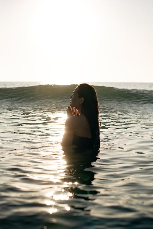 Free Side view of tranquil young female tourist with long dark hair standing in waving ocean with closed eyes and enjoying summer sunset Stock Photo