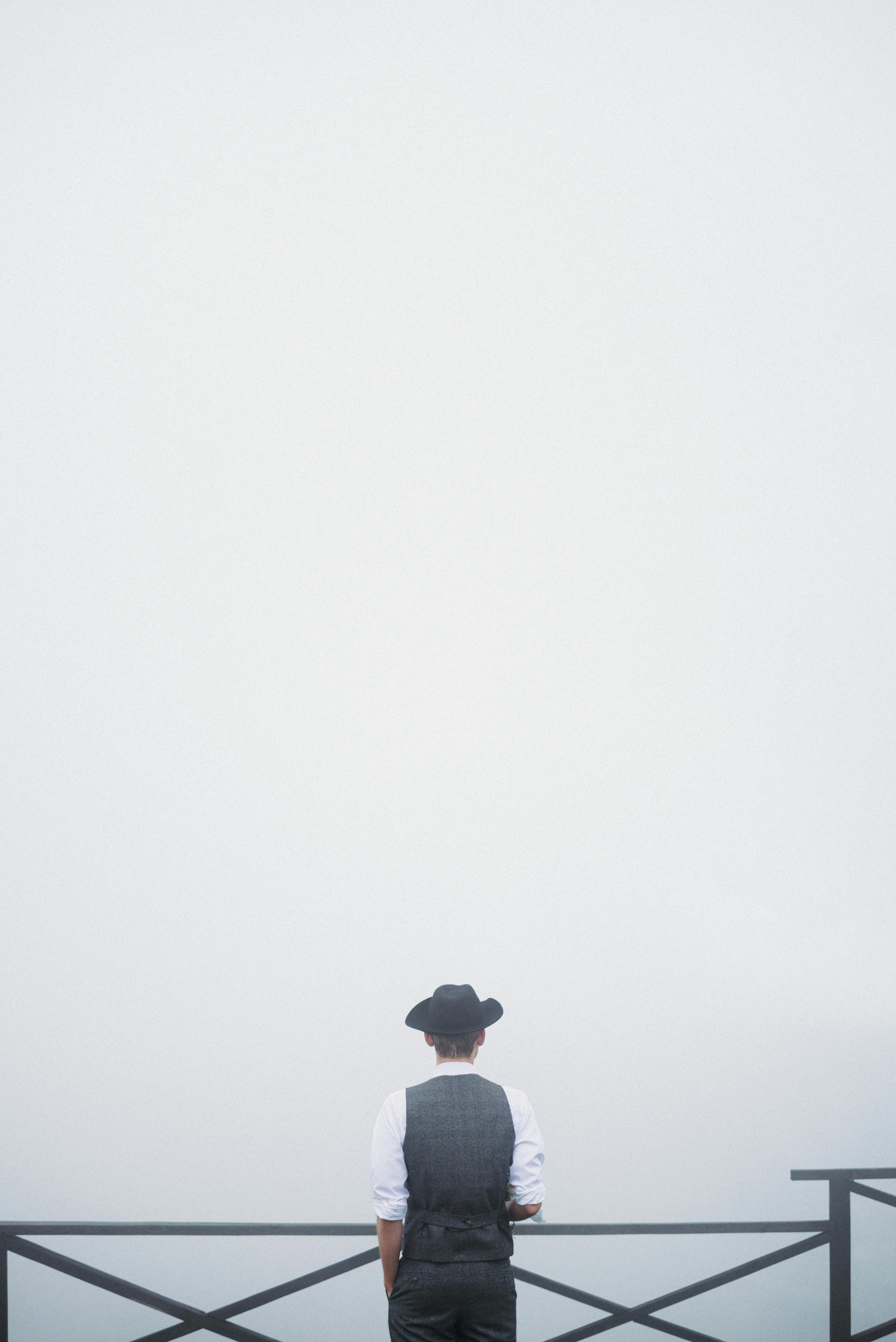 Back view of unrecognizable stylish male traveler in classic outfit and hat standing on observation platform with hand in pocket against foggy sky