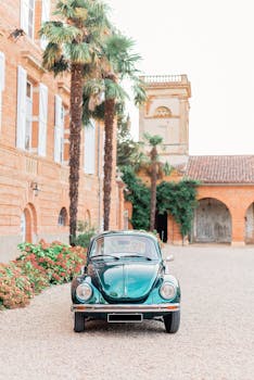 A vintage green car parked in an elegant courtyard with palm trees and historic architecture.