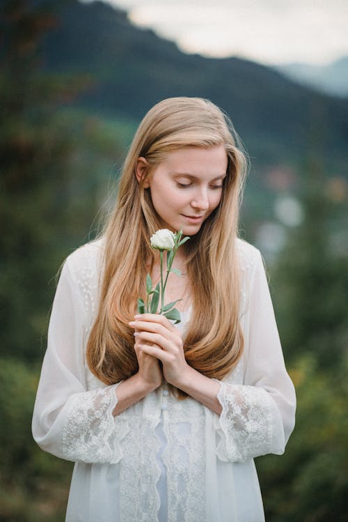 Calm young romantic female with long blond hair in white dress standing in mountainous valley with fresh flower in hand and looking down