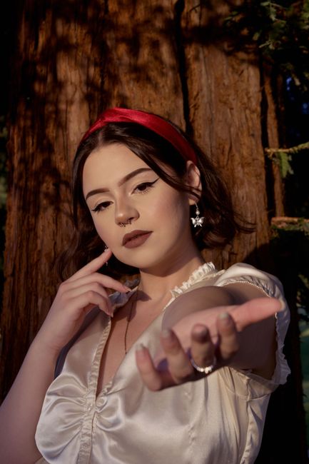 Young female with makeup in headband touching cheek while looking at camera against tree trunk in soft sunlight