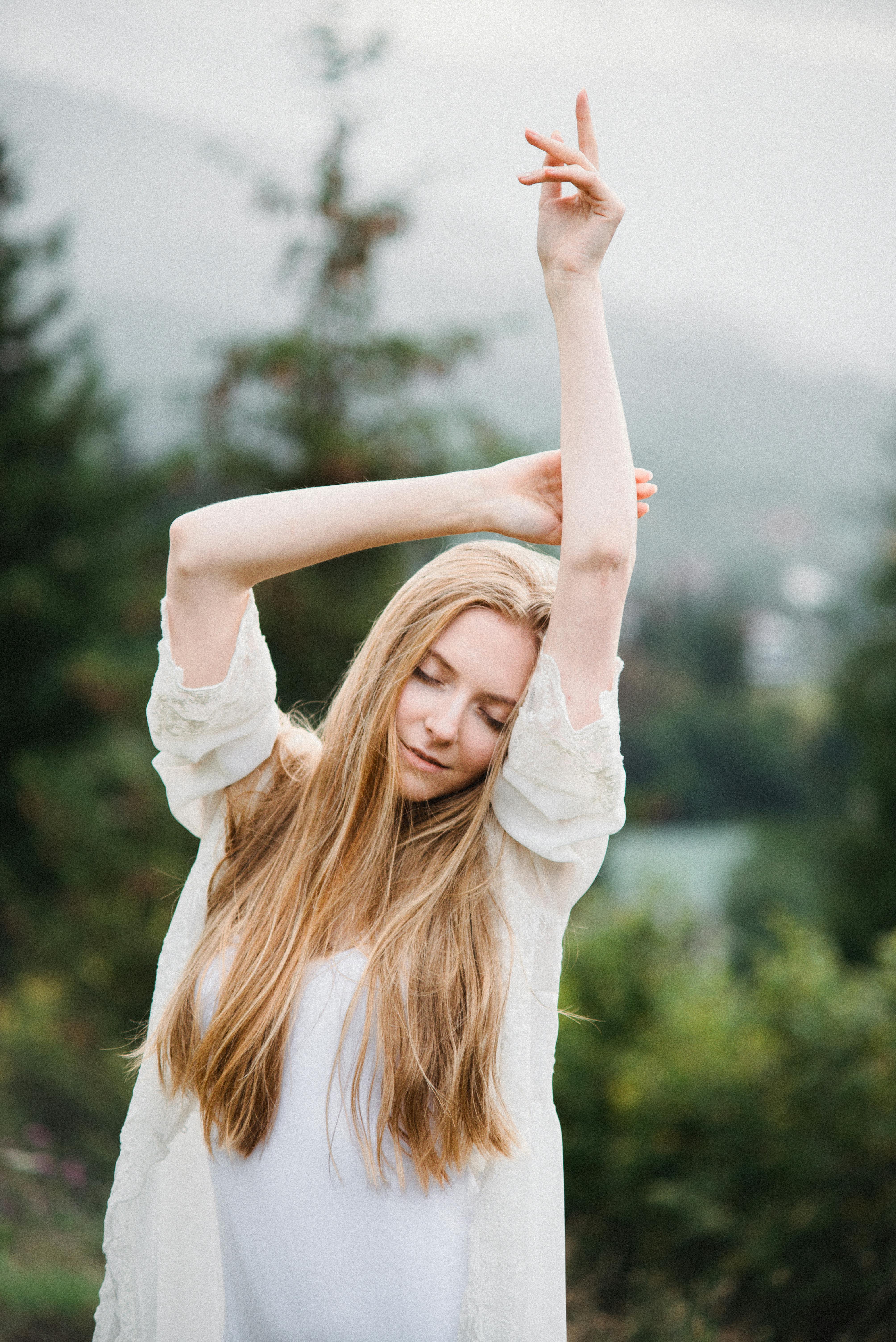 Confident young female with long blond hair in elegant white dress dancing with raised arms and closed eyes near lush green trees in mountainous valley