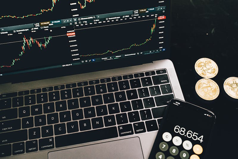 Laptop displaying cryptocurrency trading charts alongside a smartphone calculator and Bitcoin coins on a desk