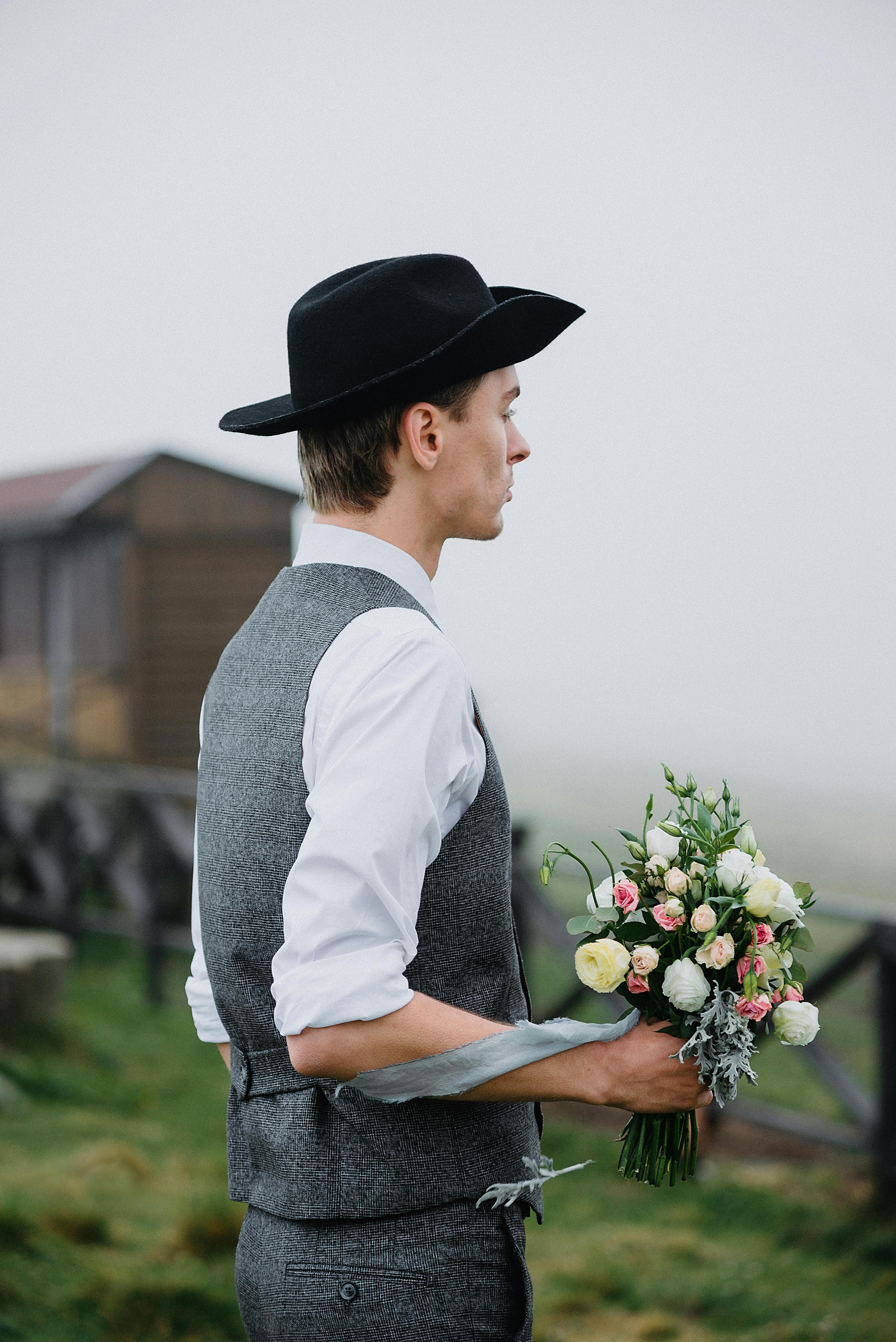 Side view of impatient young groom in elegant outfit and hat standing on grassy field with bouquet of flowers in hands and waiting for bride before wedding in countryside