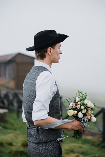 Side view of impatient young groom in elegant outfit and hat standing on grassy field with bouquet of flowers in hands and waiting for bride before wedding in countryside