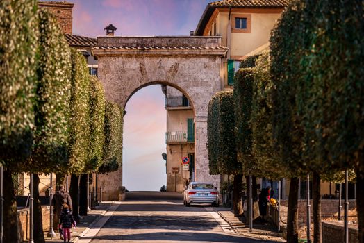 Narrow cobblestone street amidst lush green trees leading to stone arch against sunset sky in old town of Chianciano Terme