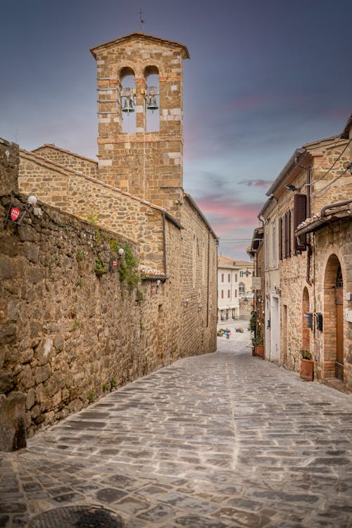 Empty cobblestone street between old house and bell tower of historic Chiesa di Sant Egidio church against cloudy sunset sky in Montalcino