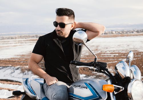 A man in sunglasses sitting on a motorbike against a scenic outdoor backdrop.