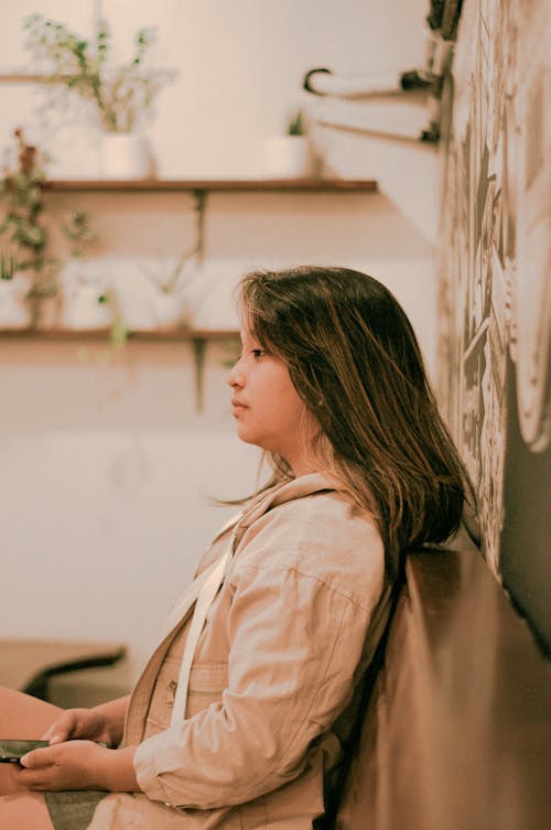 Side view of thoughtful young Asian female millennial with long hair in casual clothes resting in cozy cafe and looking away thoughtfully while messaging on mobile phone