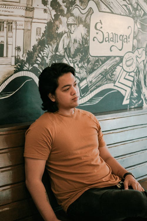 Pensive Asian male teenager with dark hair in casual outfit sitting near painted wall in cafe and looking away thoughtfully