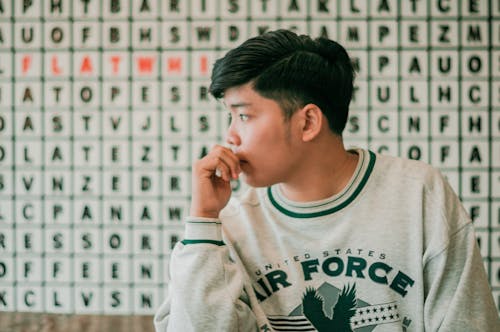 Melancholic young Asian male millennial with short dark hair in casual clothes touching lops and looking away pensively against wall with word square