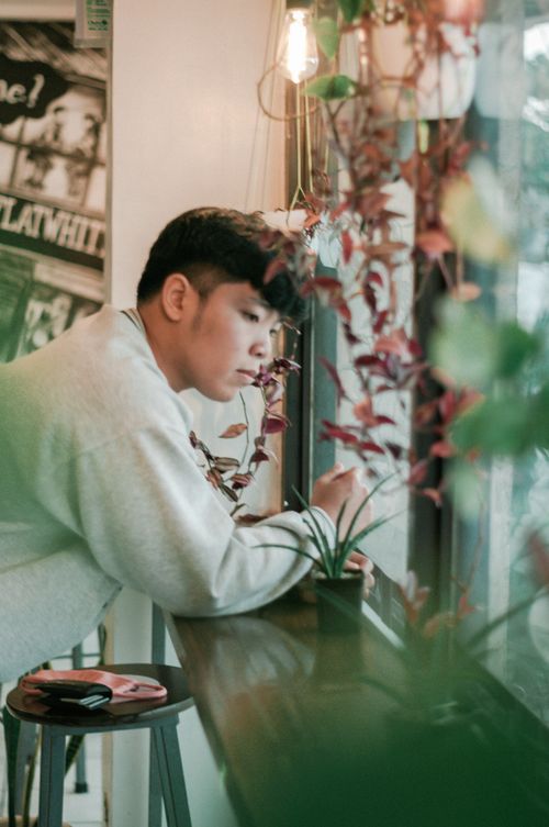 Free A man in a gray sweater leans thoughtfully on a bar table next to a window filled with plants. Stock Photo