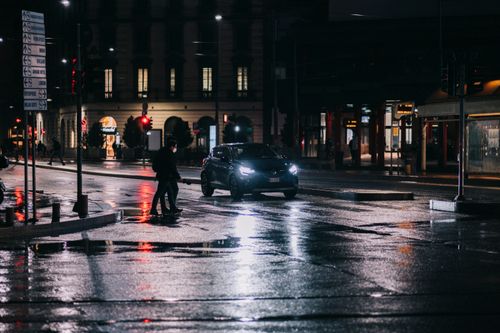 Free City street at night with car and pedestrians crossing wet road under streetlights. Stock Photo