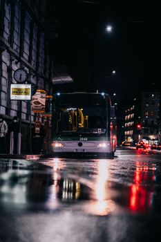 A city bus driving through rainy streets at night, illuminated by city lights and reflections.