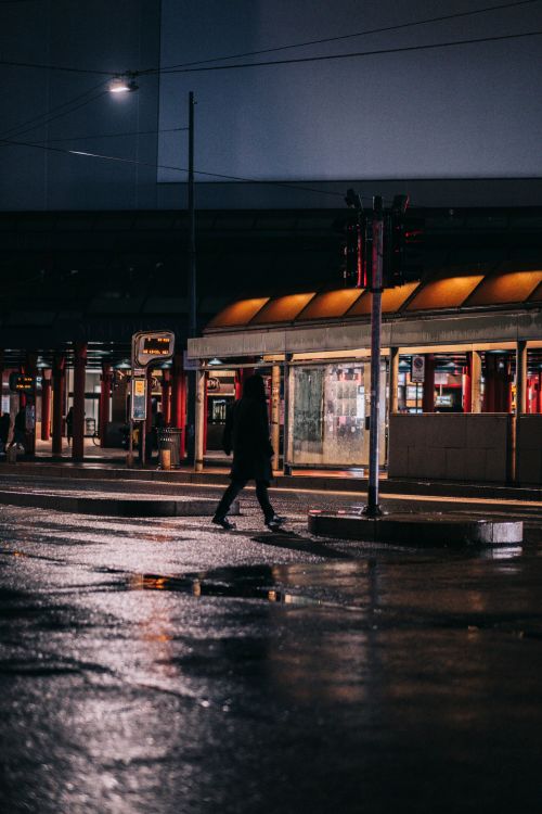 Free A person in a hoodie walks on a wet street illuminated by city lights at night. Stock Photo