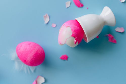 Broken Pink Egg on Egg Cup with Pieces of Eggshells on Blue Background