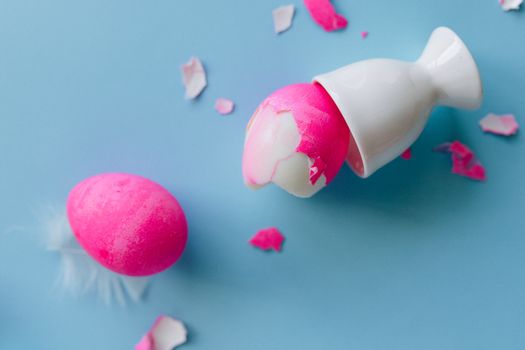 Broken Pink Egg on Egg Cup with Pieces of Eggshells on Blue Background