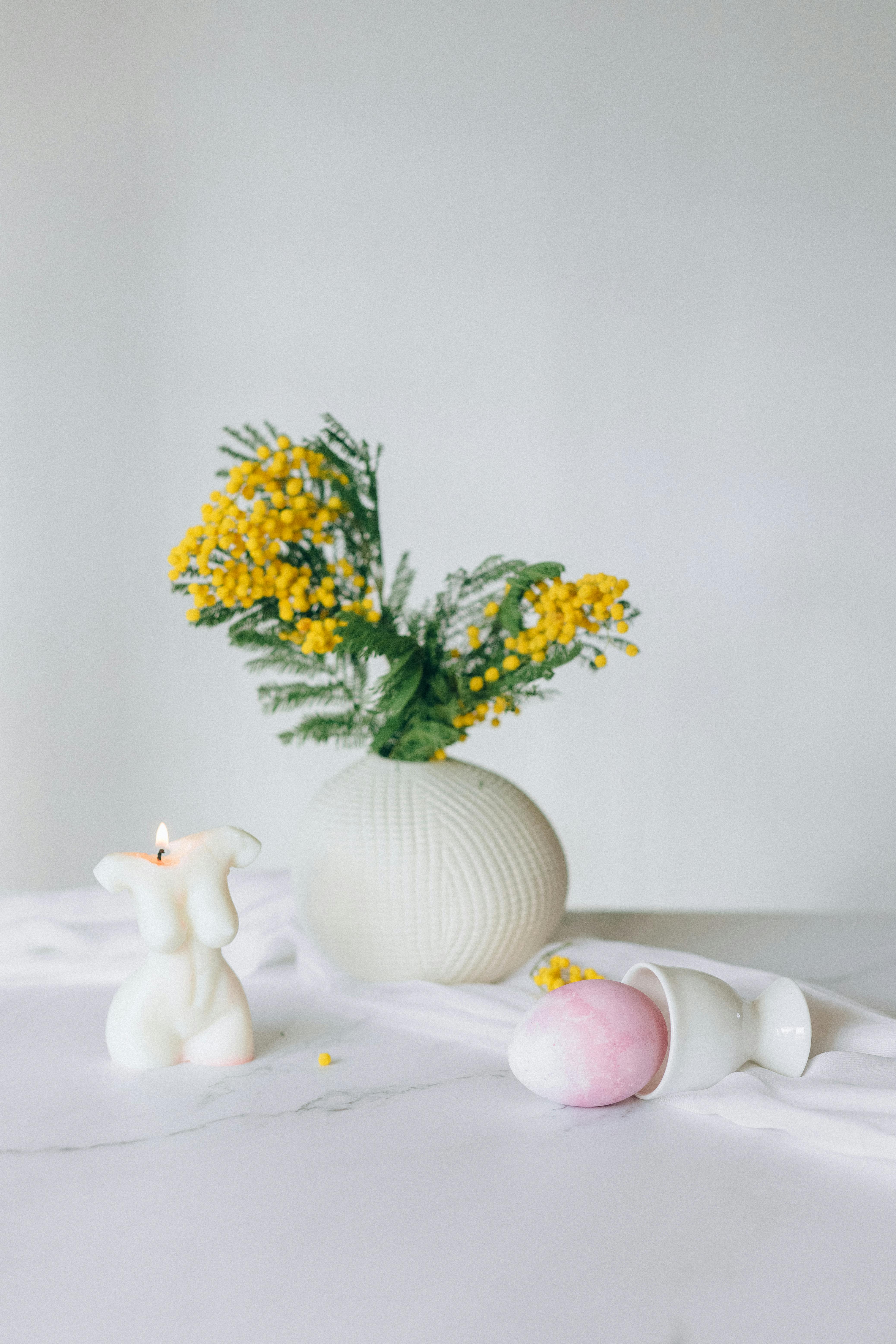 Elegant still life featuring a vase with yellow flowers, a candle, and an Easter egg on a marble surface.