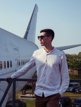 Young man in sunglasses posing stylishly by an airplane outdoors.