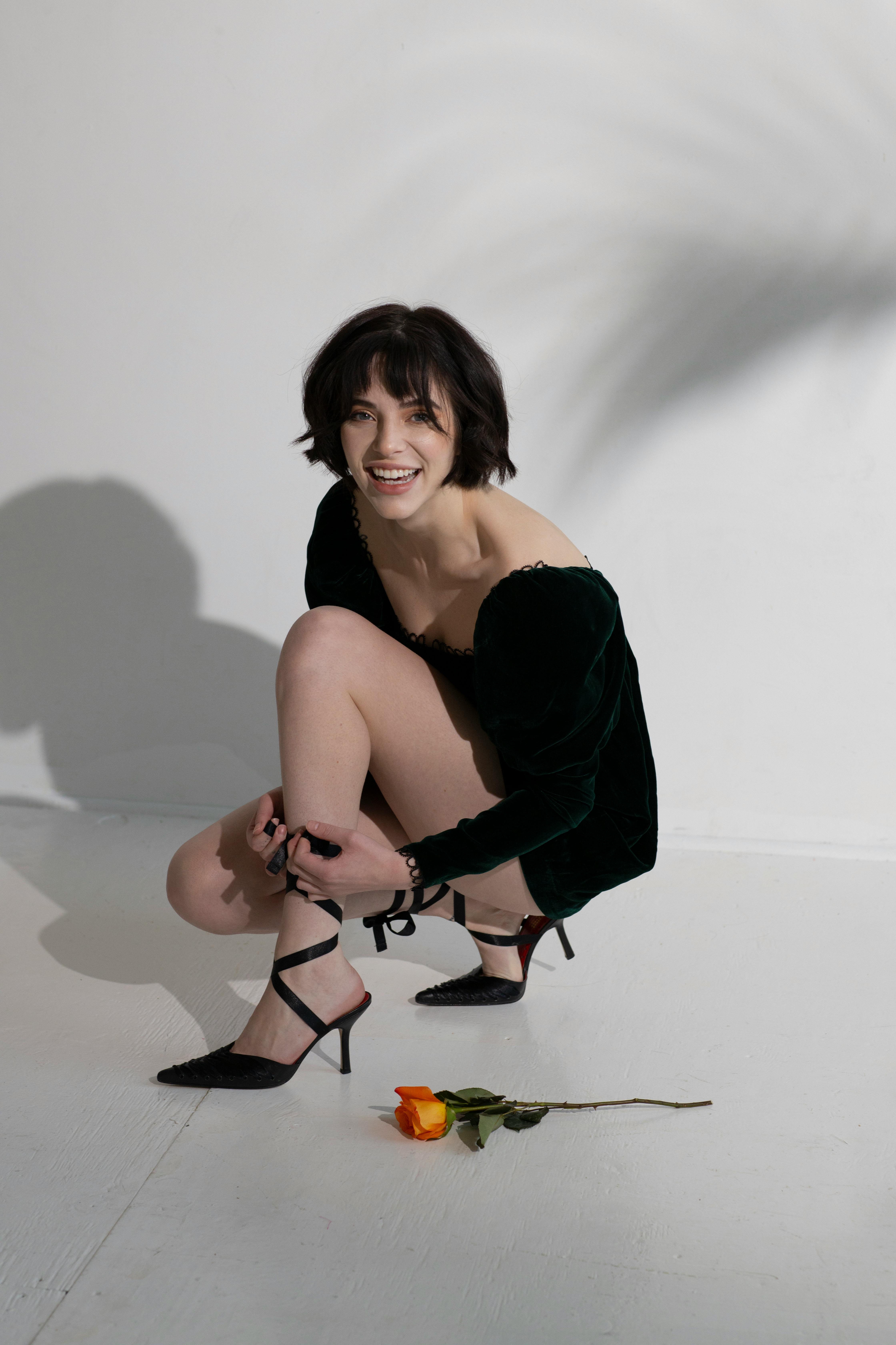 Cheerful young woman with hairstyle in stylish black dress and elegant shoes squatting down on floor near fresh rose and looking at camera against light wall with shadows