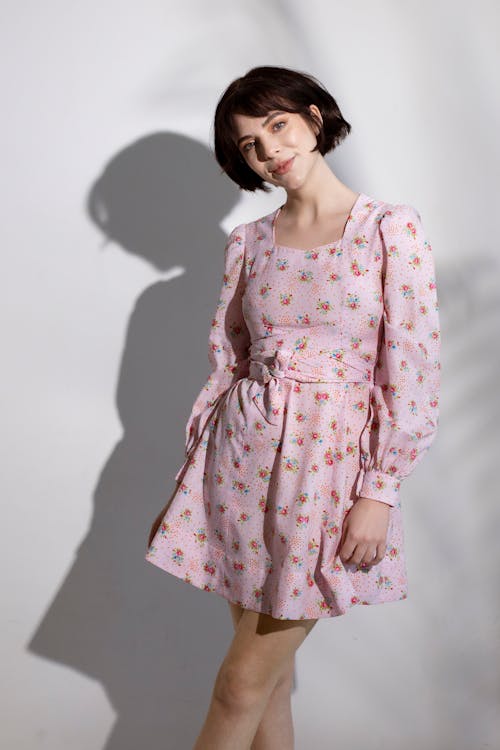 Positive woman with short brown hair in stylish dress standing against white wall and looking at camera