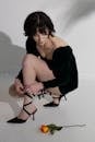 Serious female in stylish black dress tying high heels near orange rose on white floor with shades in light studio near wall
